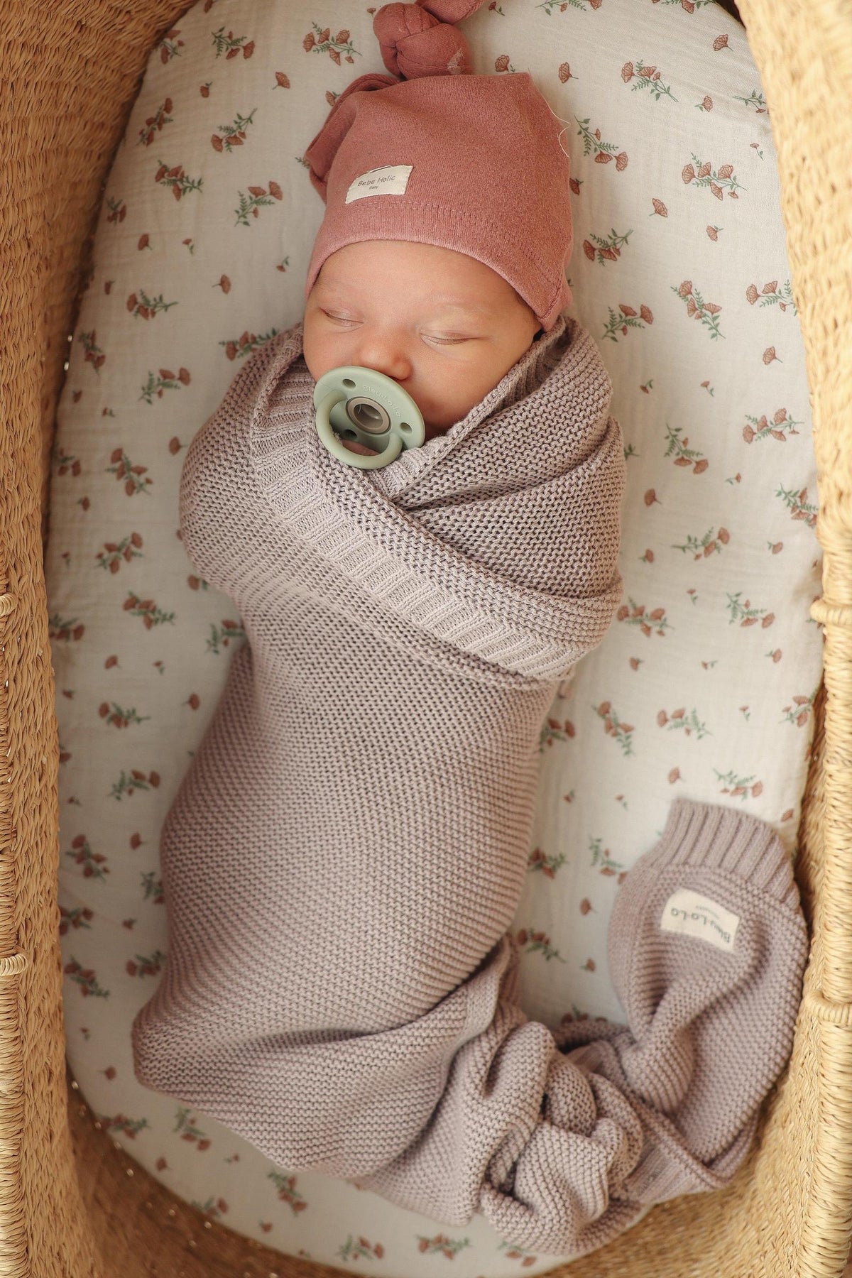 A baby is swaddled in a knitted, light purple blanket with a matching hat, lying in a woven basket.