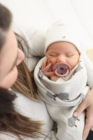 A newborn wrapped in a sheep-patterned swaddle blanket, wearing a white hat and holding a pink pacifier.