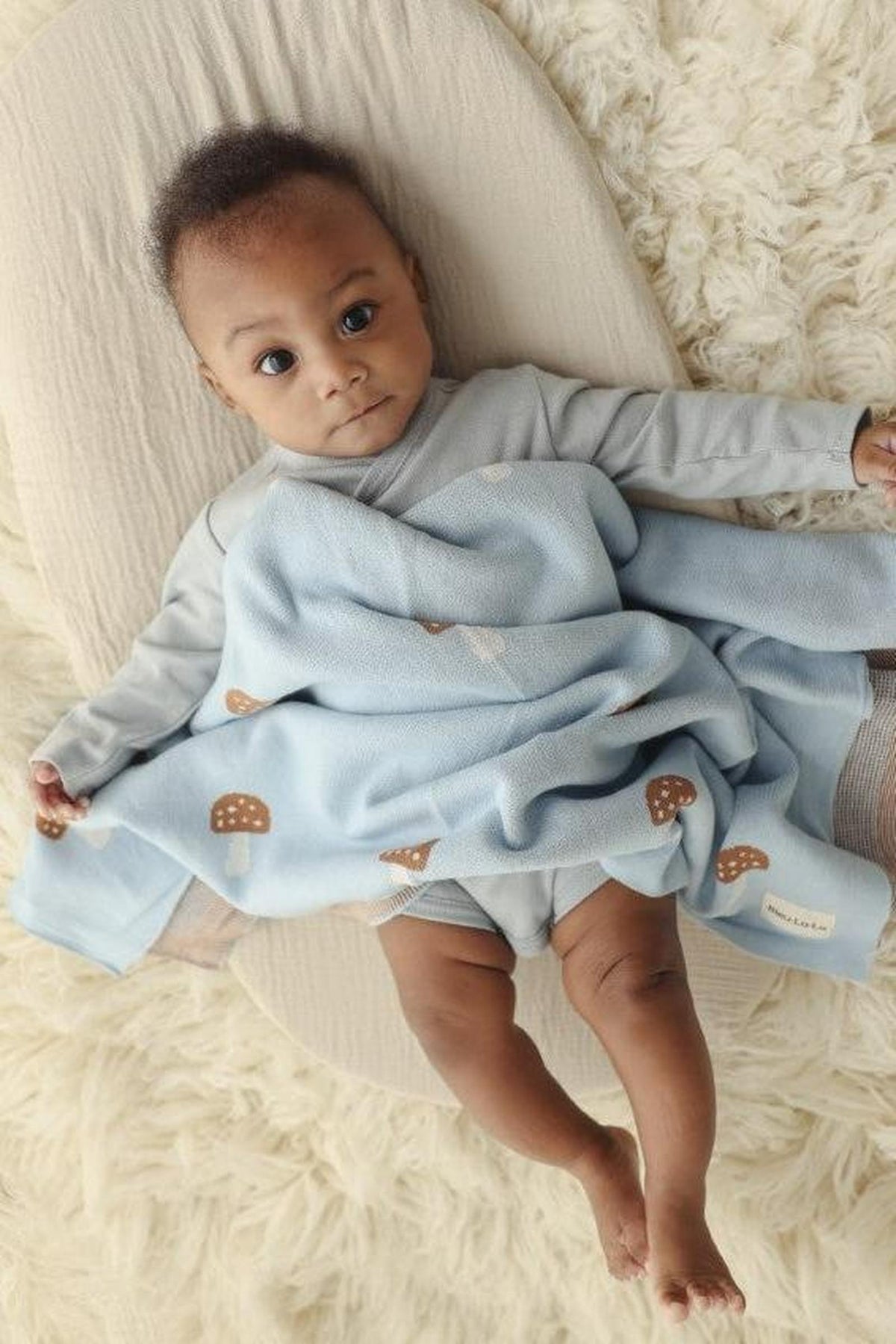 A baby is wrapped in a light blue blanket with mushroom patterns, lying on a soft beige surface.