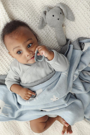 Baby wrapped in a blue knit blanket with llama design, holding a teether, next to a gray plush toy.
