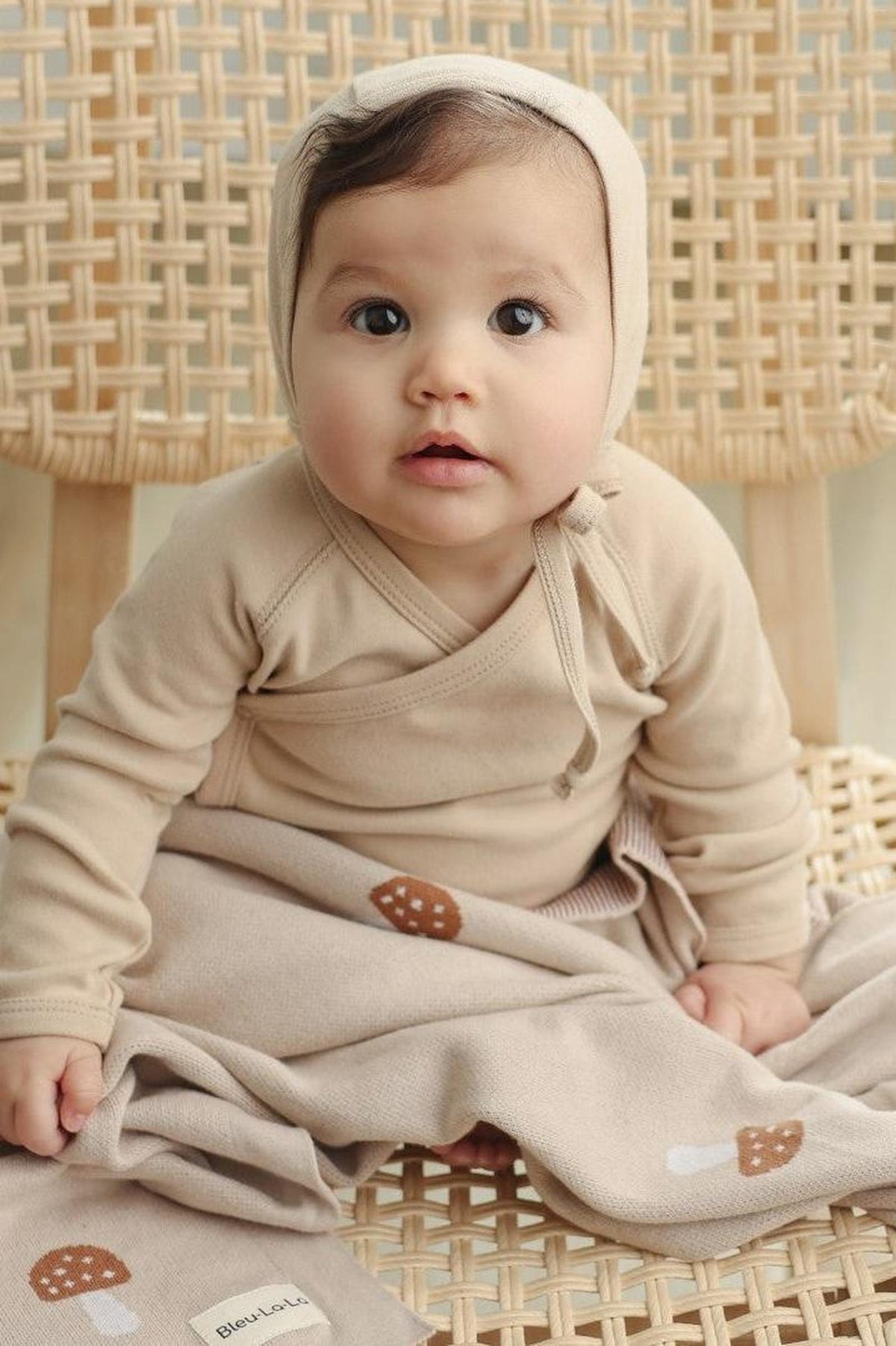 A baby in a beige outfit sits on a woven chair, wrapped in a mushroom-patterned blanket.