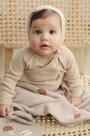 A baby in a beige outfit sits on a woven chair, wrapped in a mushroom-patterned blanket.