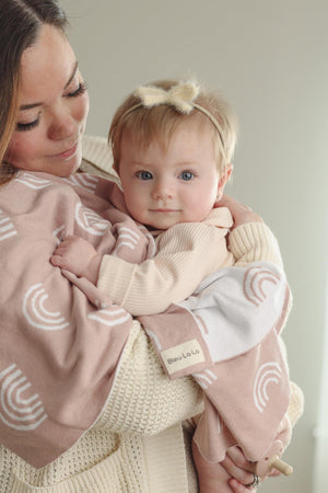 A baby with a soft headband is wrapped in a pink swaddle blanket featuring white rainbow patterns.