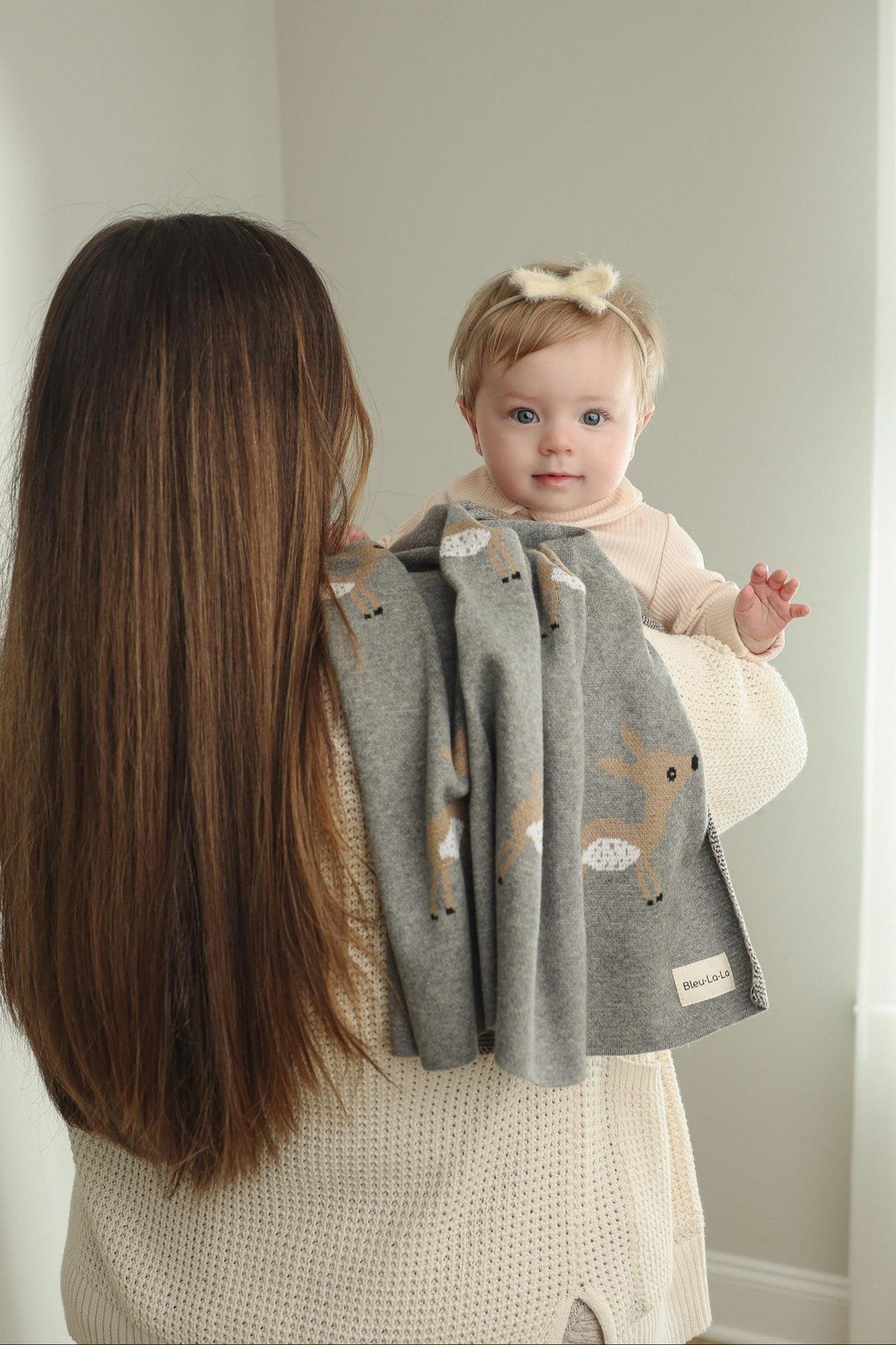 A woman holds a baby wrapped in a gray deer-patterned knit blanket, with soft natural lighting.