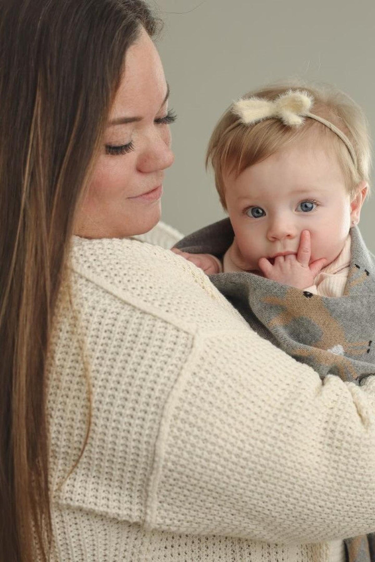 A baby wrapped in a grey blanket with deer design is held by a woman in a cream sweater.