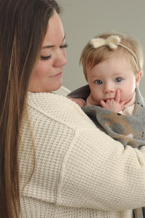 A baby wrapped in a grey blanket with deer design is held by a woman in a cream sweater.