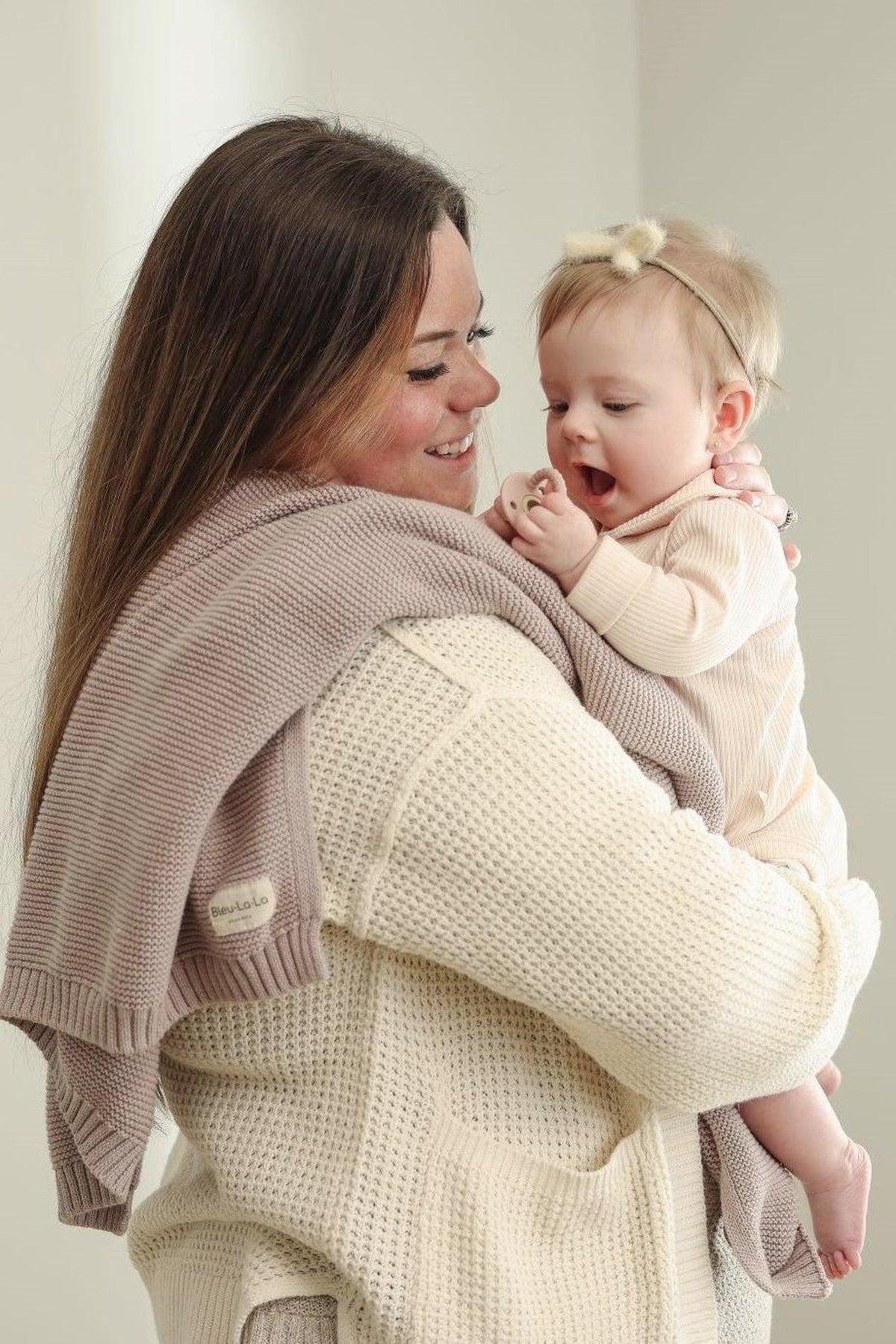 A woman in a cream sweater holds a baby with a beige bow headband, wrapped in a taupe knit blanket.