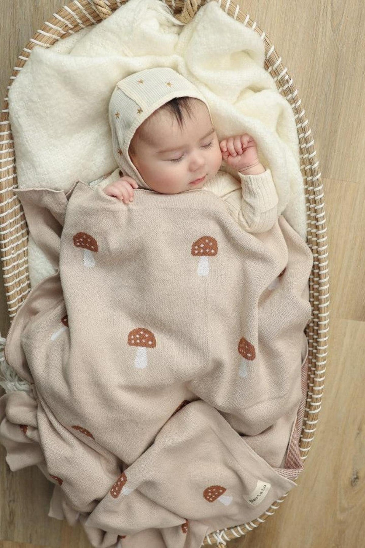 A baby sleeps peacefully in a basket, covered with a beige mushroom-patterned knit blanket.