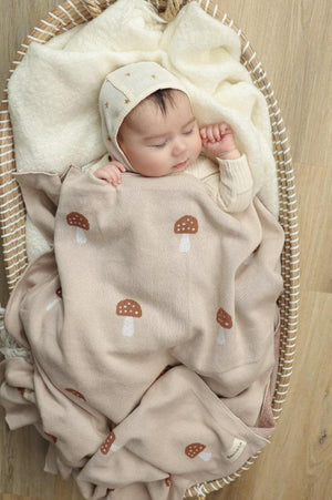 A baby sleeps peacefully in a basket, covered with a beige mushroom-patterned knit blanket.
