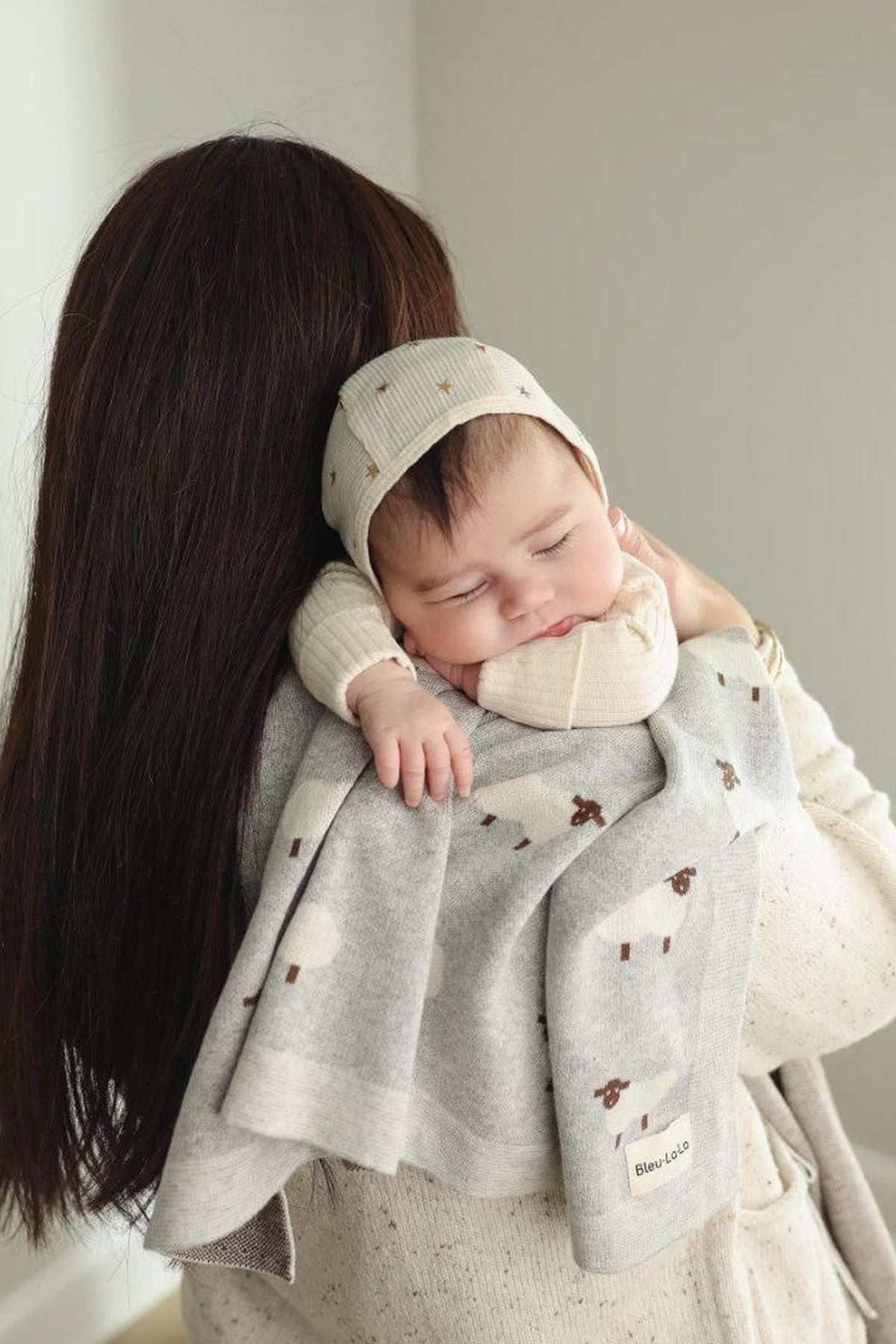 A sleeping baby rests on a womans shoulder, wrapped in a gray sheep-patterned knit blanket.
