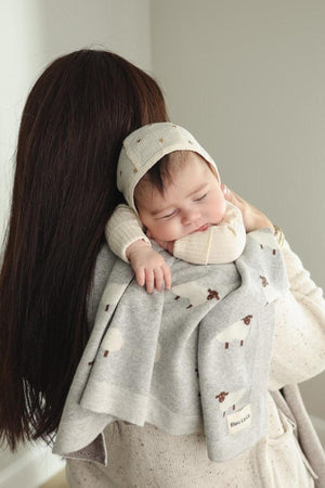 A sleeping baby rests on a womans shoulder, wrapped in a gray sheep-patterned knit blanket.
