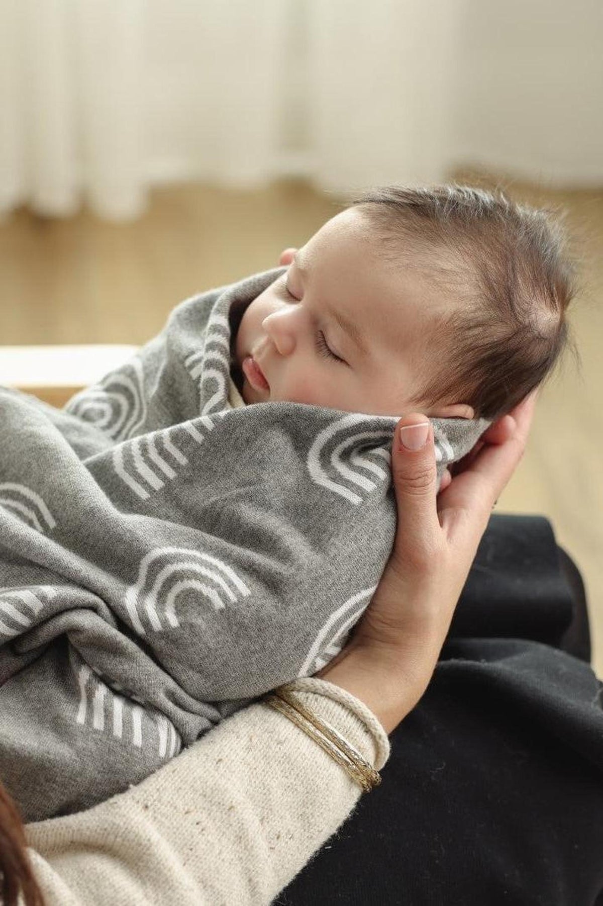 A baby wrapped in a gray swaddle blanket with white rainbow patterns, held gently by a person.