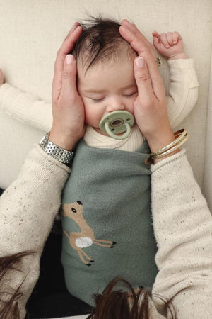 A baby with a pacifier is wrapped in a green swaddle featuring a deer design, cradled by hands.