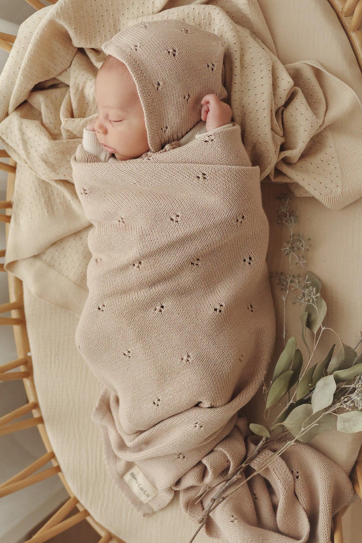 A baby is swaddled in a beige, pointelle knit blanket, lying in a wicker basket with eucalyptus leaves.