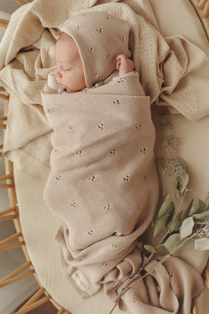 A baby is swaddled in a beige, pointelle knit blanket, lying in a wicker basket with eucalyptus leaves.