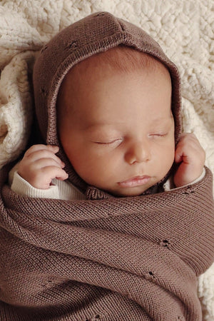 A sleeping baby wrapped in a brown knit swaddle blanket, resting on a textured cream background.