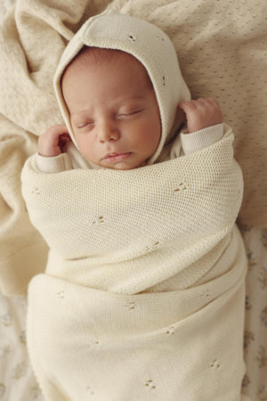 A newborn baby peacefully sleeps, wrapped in a cream-colored organic pointelle swaddle blanket with a matching hat.