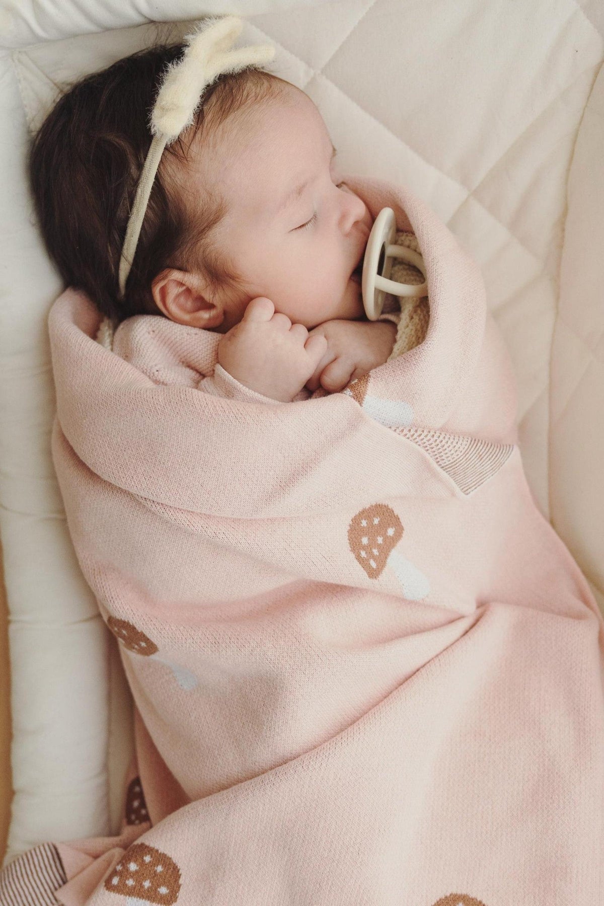 A baby sleeps peacefully wrapped in a pink mushroom-patterned swaddle blanket, wearing a soft headband with a pacifier.