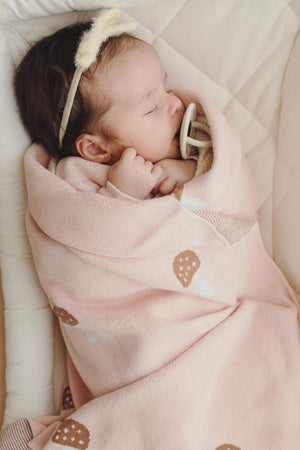 A baby sleeps peacefully wrapped in a pink mushroom-patterned swaddle blanket, wearing a soft headband with a pacifier.