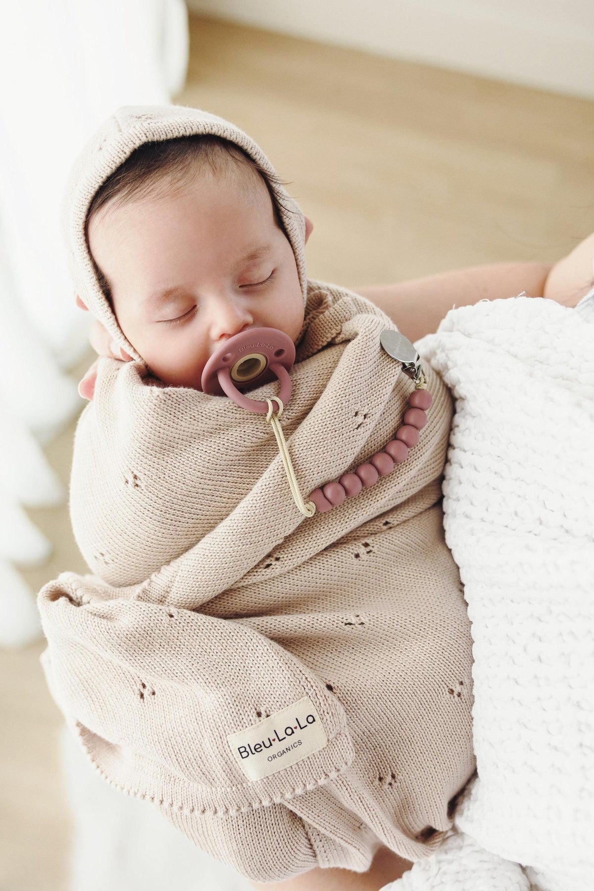A sleeping baby wrapped in a beige organic pointelle blanket with a matching hat and pacifier clip.