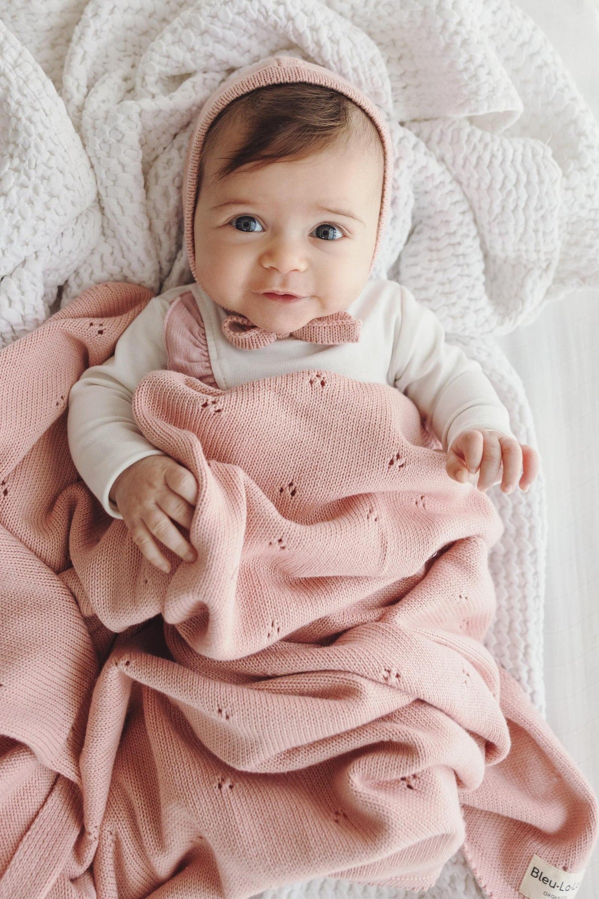 A baby wrapped in a pink pointelle knit swaddle blanket, lying on a textured white blanket.