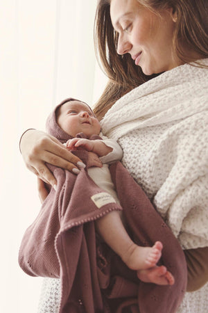 A mother holds her baby wrapped in a mauve pointelle blanket with a matching hat, both smiling warmly.