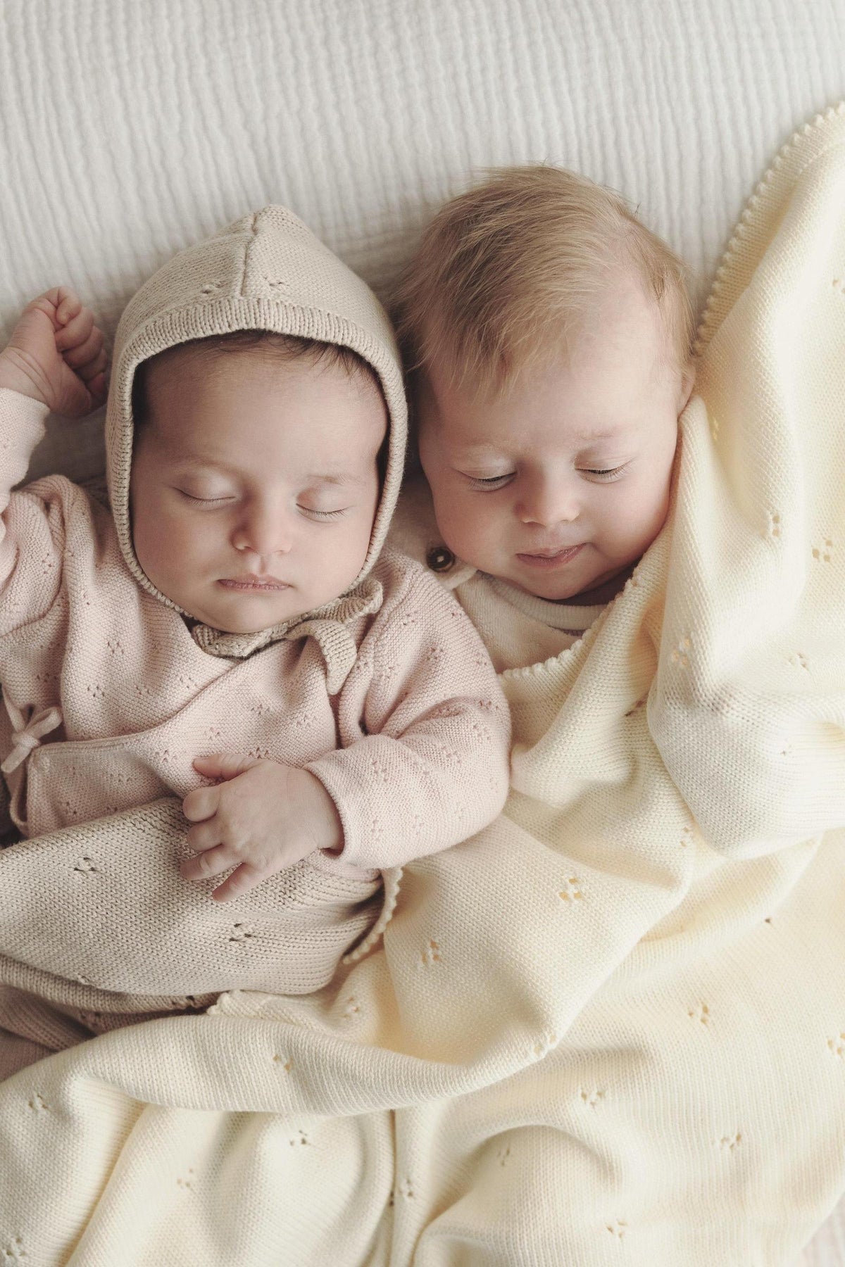 Two babies sleeping on a soft, cream pointelle blanket, one wearing a matching beige knit hat.