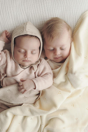 Two babies sleeping on a soft, cream pointelle blanket, one wearing a matching beige knit hat.