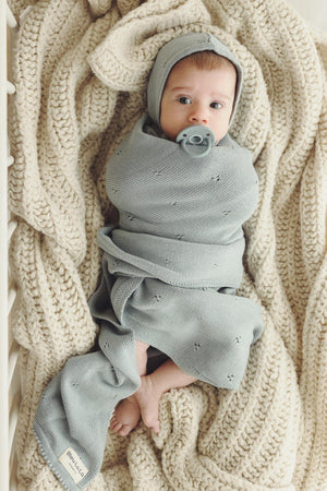 A baby wrapped in a light gray knit swaddle blanket with a matching hat, lying on a cream knitted blanket.