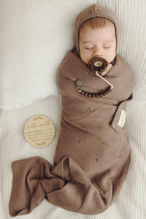 A baby sleeps wrapped in a brown pointelle swaddle blanket, wearing a matching hat, with a pacifier.