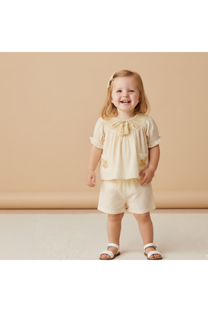 A smiling toddler wears a soft, butter-yellow organic smocked top with delicate embroidery and matching shorts. The outfit features a lightweight, breathable fabric, perfect for sunny day outings.