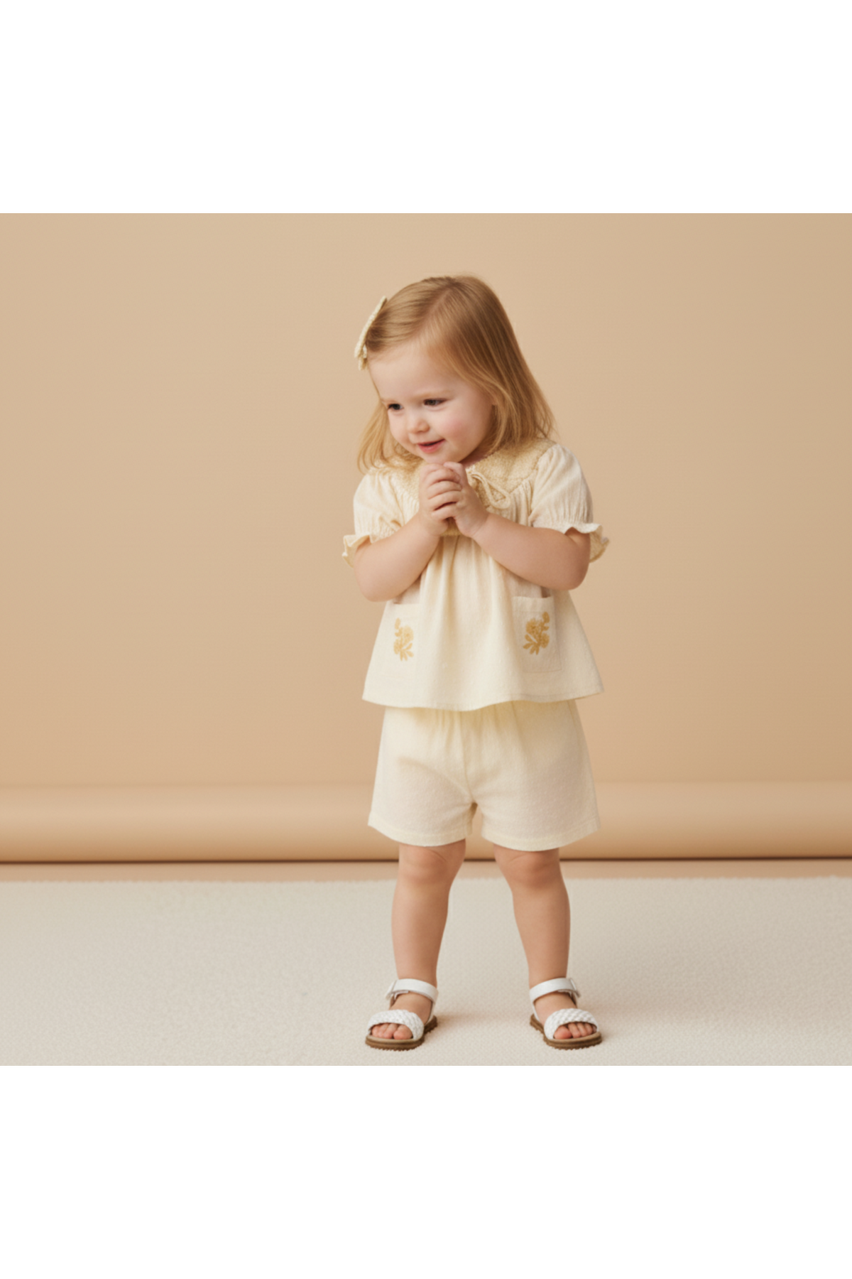 A young child smiles while wearing a soft, organic cotton smocked top and shorts set in butter yellow, featuring delicate embroidery. The outfit is perfect for sunny day outings.