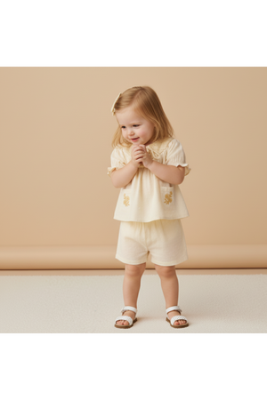 A young child smiles while wearing a soft, organic cotton smocked top and shorts set in butter yellow, featuring delicate embroidery. The outfit is perfect for sunny day outings.