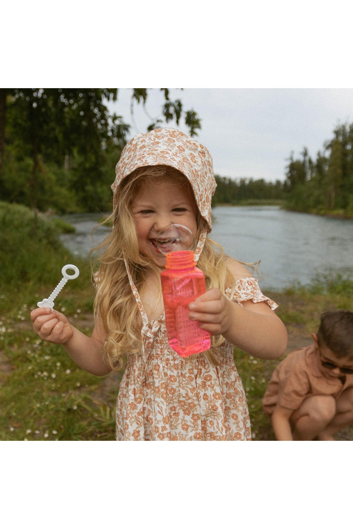 A smiling child wearing a floral sundress and a sun hat holds a bottle of pink bubble solution by a riverbank, surrounded by greenery and a serene landscape.