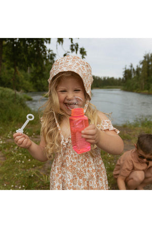 A smiling child wearing a floral sundress and a sun hat holds a bottle of pink bubble solution by a riverbank, surrounded by greenery and a serene landscape.