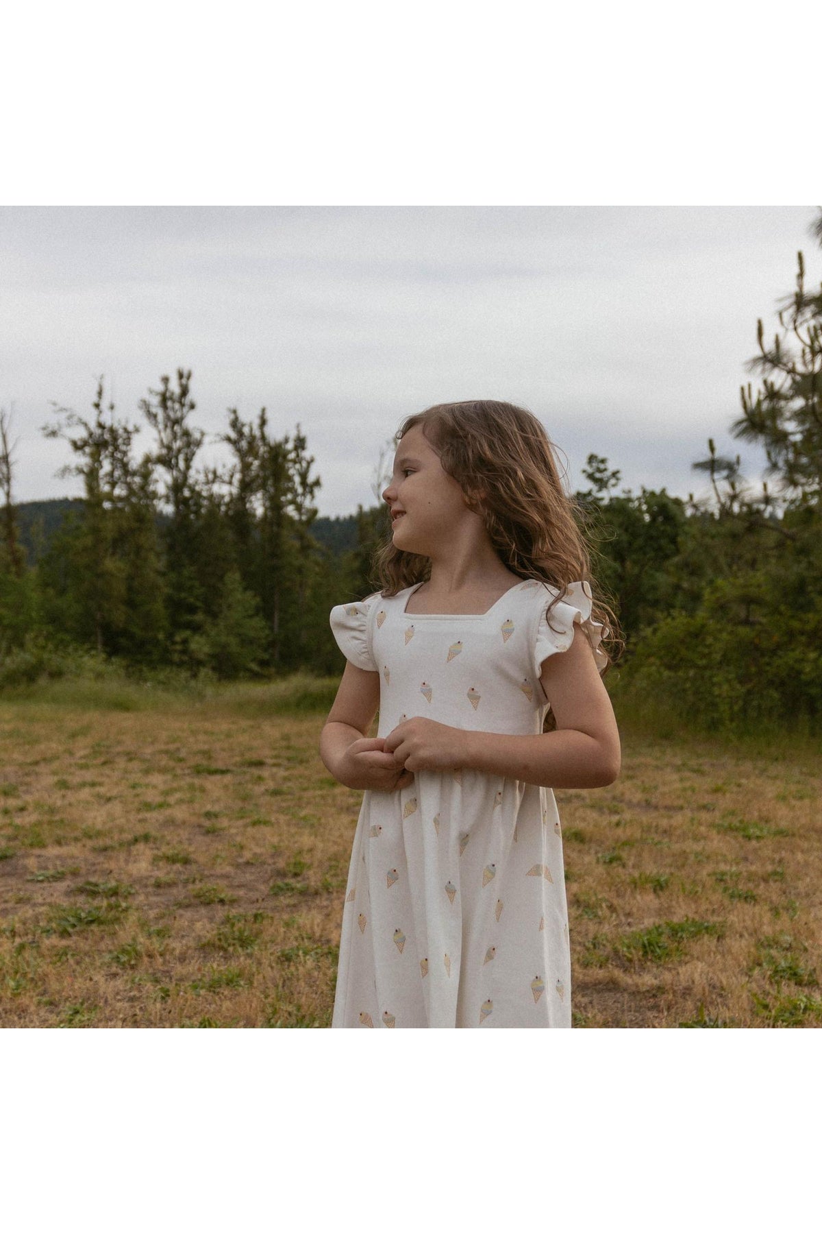 A young girl stands in a grassy area, wearing a cream-colored dress adorned with an ice cream print.
