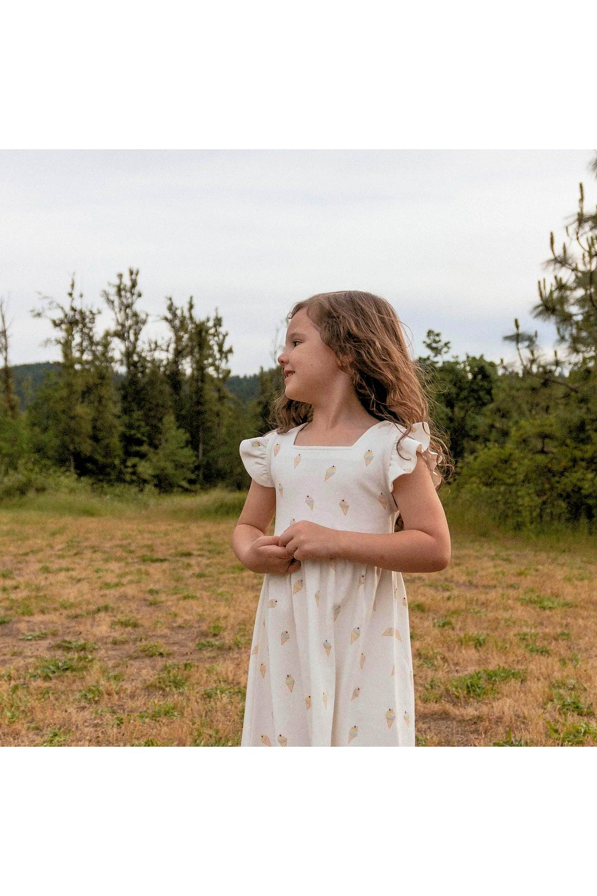A young girl stands in a grassy field, wearing a light-colored dress adorned with an ice cream print.