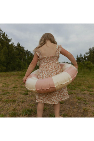 A young girl wearing a floral smocked sundress holds a large pink and cream inflatable float. She stands in a grassy area surrounded by trees under a cloudy sky.