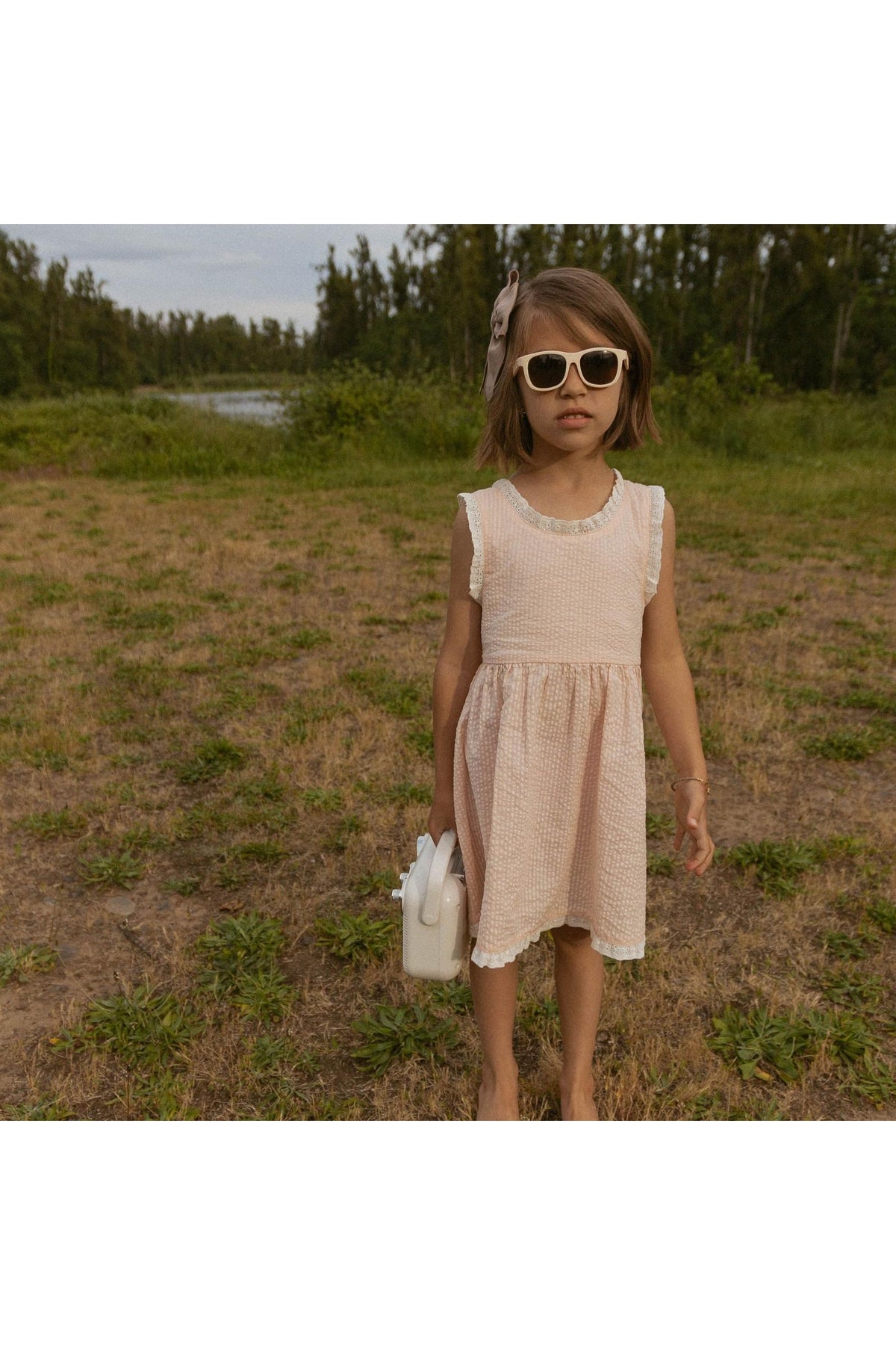 A young girl stands outdoors wearing a blush organic cotton dress with criss-cross straps, paired with oversized sunglasses and holding a small bag.