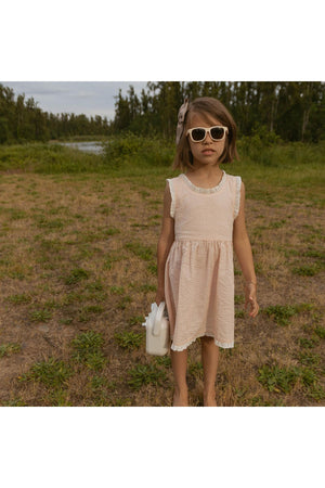 A young girl stands outdoors wearing a blush organic cotton dress with criss-cross straps, paired with oversized sunglasses and holding a small bag.