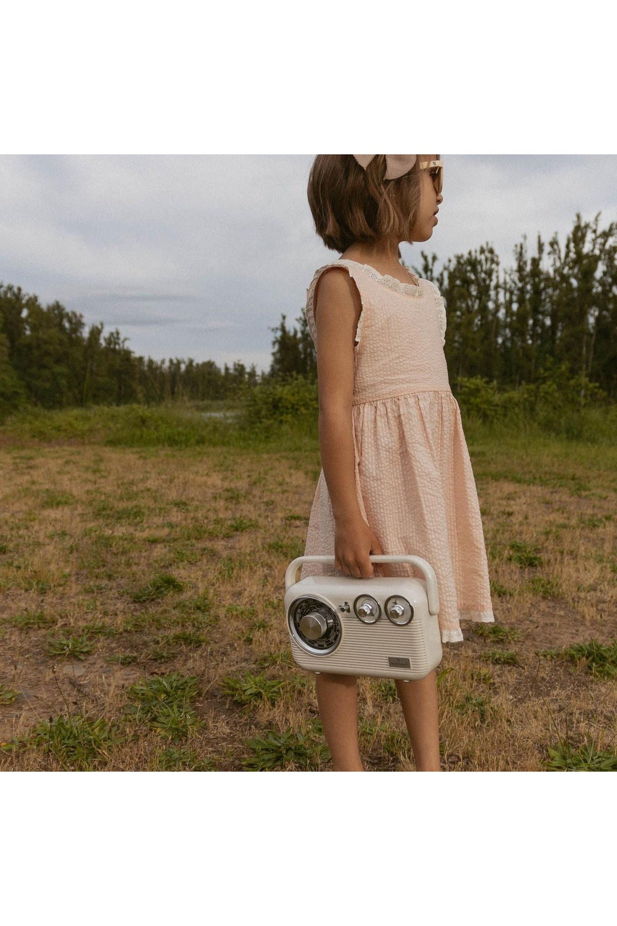 A young girl stands outdoors, wearing a blush-colored dress with criss-cross straps. She holds a vintage-style radio, surrounded by lush greenery and an overcast sky.