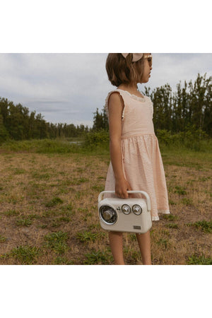 A young girl stands outdoors, wearing a blush-colored dress with criss-cross straps. She holds a vintage-style radio, surrounded by lush greenery and an overcast sky.