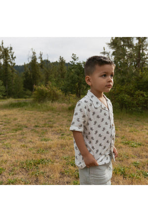 A young boy stands outdoors in a grassy area, wearing a short-sleeved, patterned shirt and light shorts. The outfit features organic materials, reflecting a sustainable and stylish summer look.