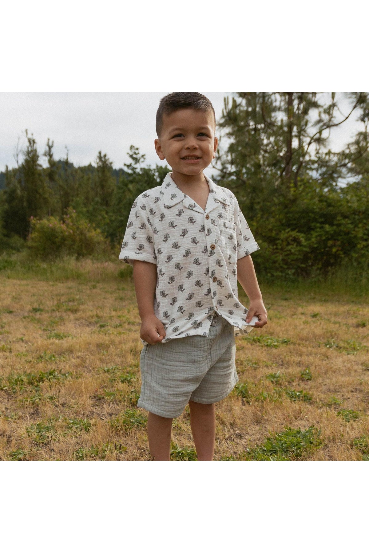 A young boy smiles while wearing a short-sleeved, white shirt with a subtle leaf pattern and light gray shorts, standing in a grassy area surrounded by trees.