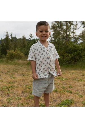 A young boy smiles while wearing a short-sleeved, white shirt with a subtle leaf pattern and light gray shorts, standing in a grassy area surrounded by trees.