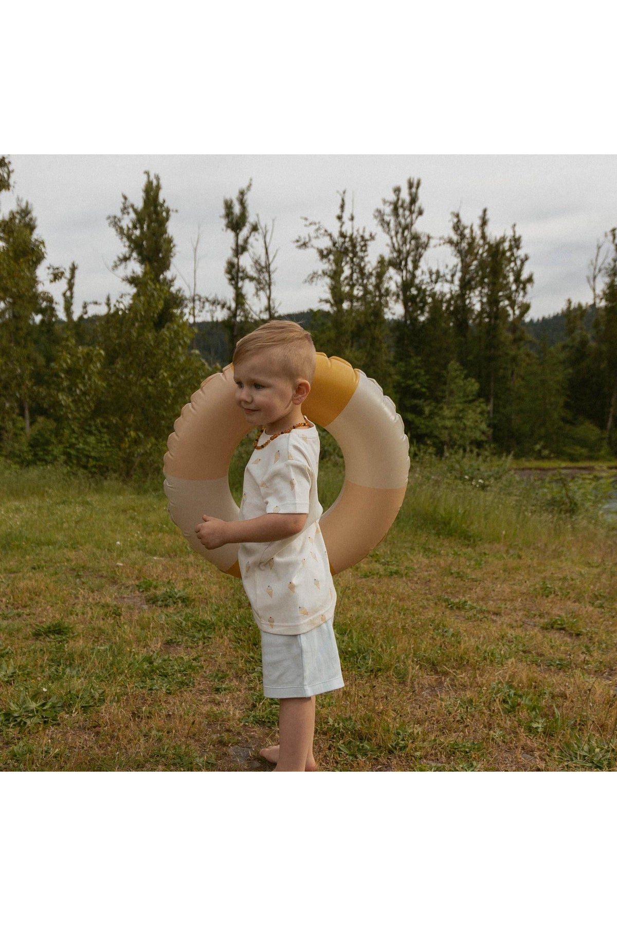 A young child stands outdoors, holding a large, soft-colored inflatable ring. They are dressed in a light-colored shirt and shorts, surrounded by greenery and a calm landscape.