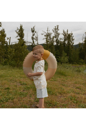 A young child stands outdoors, holding a large, soft-colored inflatable ring. They are dressed in a light-colored shirt and shorts, surrounded by greenery and a calm landscape.