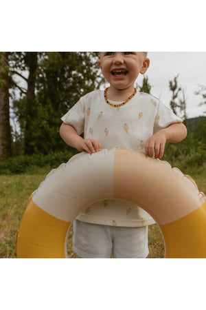 A smiling child wears a cream tee with an ice cream print and white shorts, holding a yellow and beige inflatable ring in a grassy outdoor setting surrounded by trees.