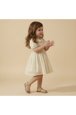 A young girl stands playfully in a soft butter yellow organic cotton dress with a smocked collar. The dress features a classic silhouette, perfect for casual outings or special occasions.