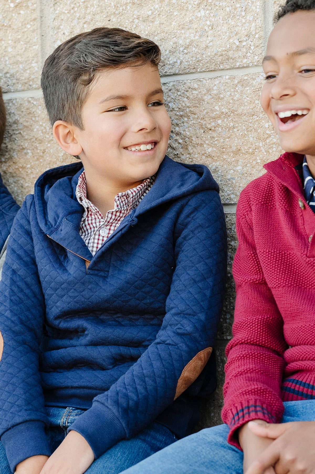 Two boys smiling, one wearing a navy quilted hooded sweatshirt with elbow patches, sitting against a textured wall.